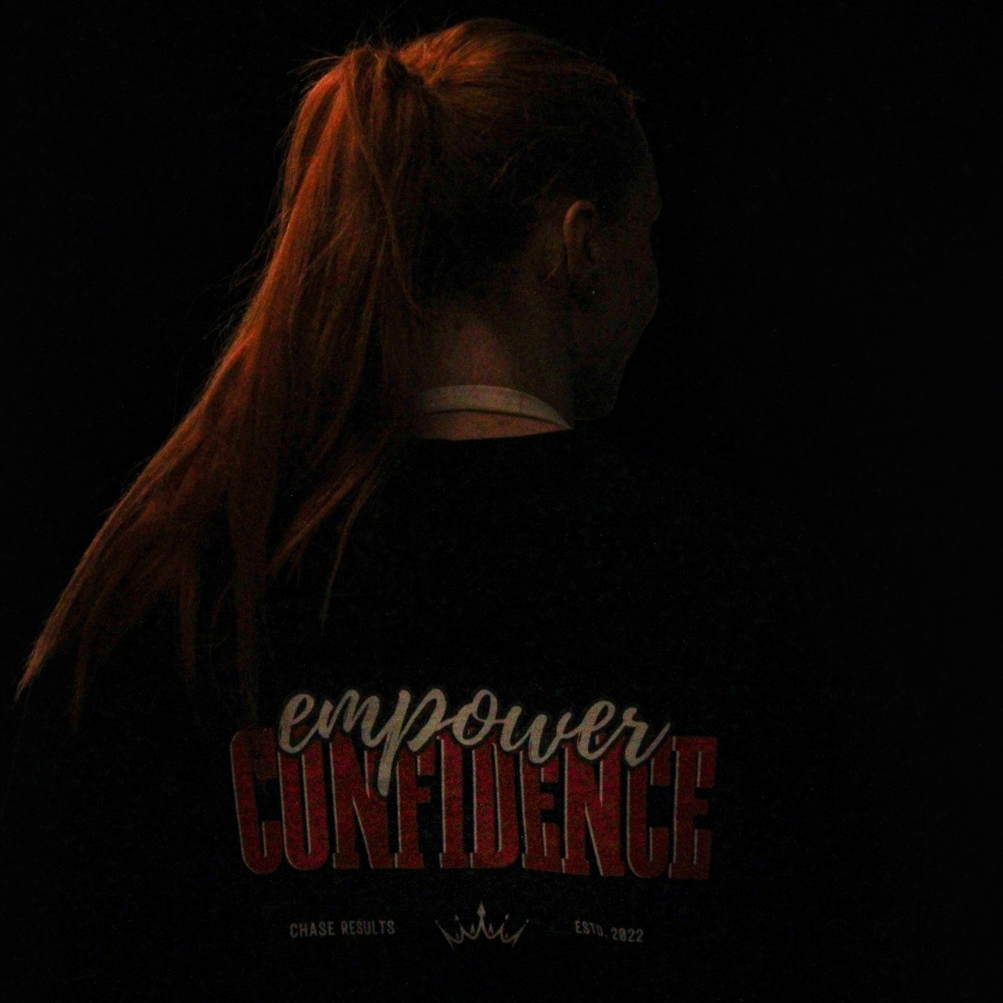 one individual wearing Chase Results Clothing's Empower Confidence T-Shirt Black Edition pose in dimly lit Great Yarmouth, one facing forward with long orange hair, the other turned away, capturing the brands dynamic spirit.