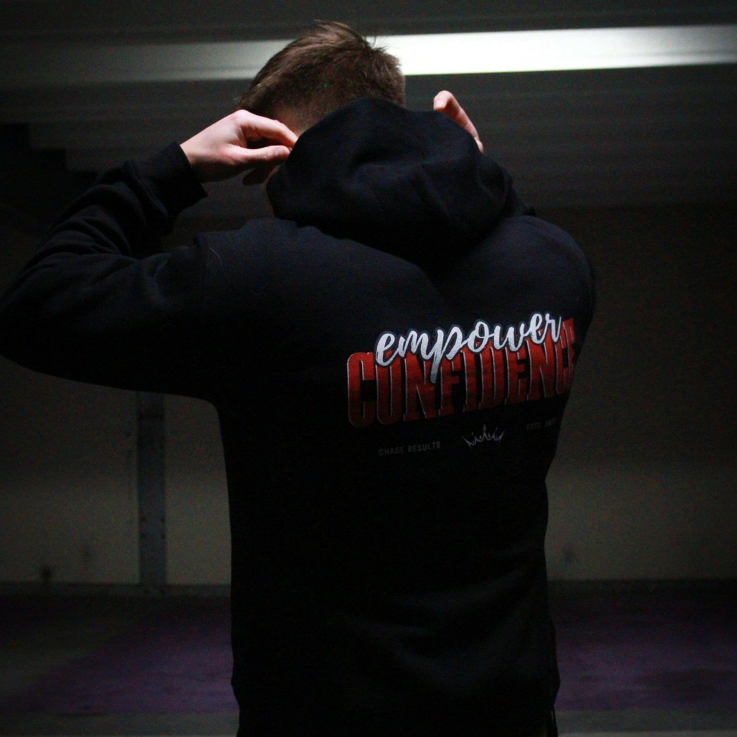 a person pulls up the hood of their limited edition Empower Confidence Black Hoodie by Chase Results Clothing against a textured gray and black wall backdrop, casting soft shadows on their face and exuding empowerment.