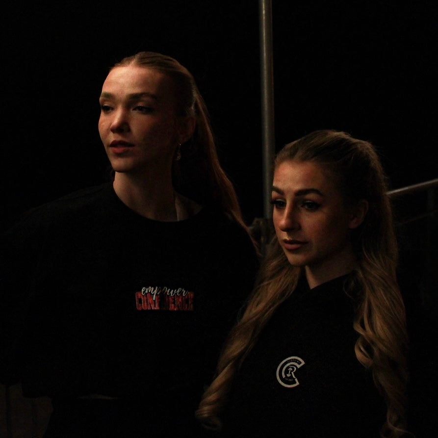 Two individuals wearing Chase Results Clothings Empower Confidence T-Shirt Black Edition pose in dimly lit Norwich, one facing forward with long orange hair, the other turned away, capturing the brands dynamic spirit.