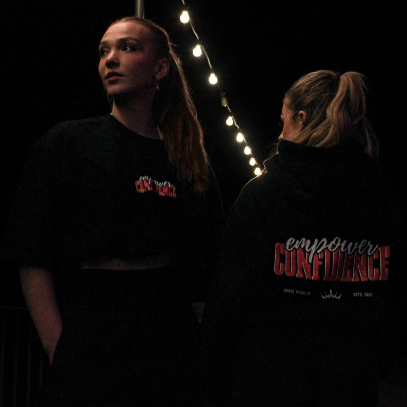 Two individuals wearing Chase Results Clothings Empower Confidence T-Shirt Black Edition pose in dimly lit Great Yarmouth, one facing forward with long orange hair, the other turned away, capturing the brands dynamic spirit.