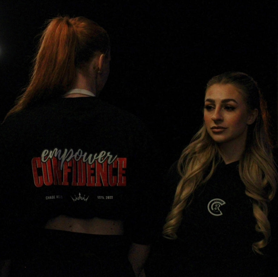 Two individuals wearing Chase Results Clothings Empower Confidence T-Shirt Black Edition pose in dimly lit Great Yarmouth, one facing forward with long orange hair, the other turned away, capturing the brands dynamic spirit.