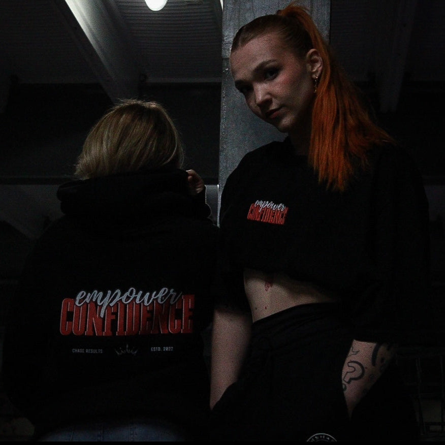 Two individuals wearing Chase Results Clothings Empower Confidence T-Shirt Black Edition pose in dimly lit Great Yarmouth, one facing forward with long orange hair, the other turned away, capturing the brands dynamic spirit.