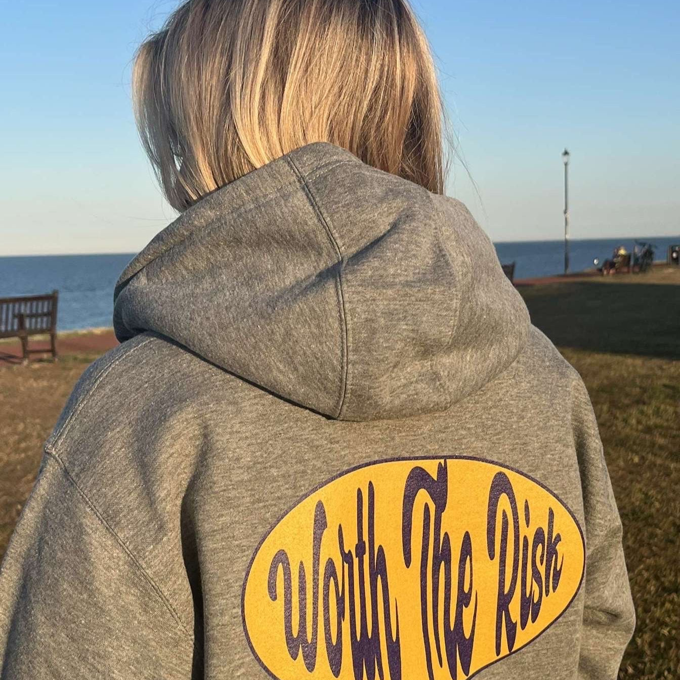Person wearing a gray hoodie with 'Worth The Risk' text at a beach.