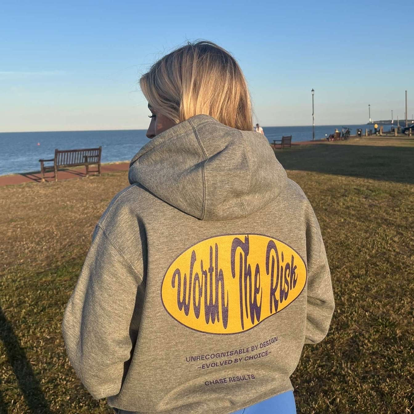 Person wearing a gray hoodie with 'Worth The Risk' text at a park by the water.