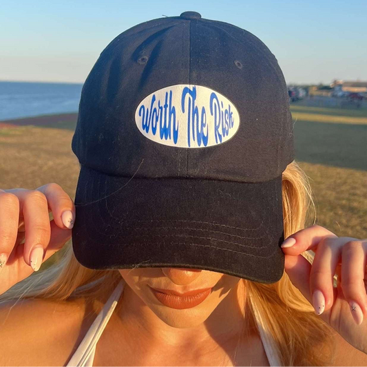 Person wearing a black cap with 'Worth The Risk' text at the beach