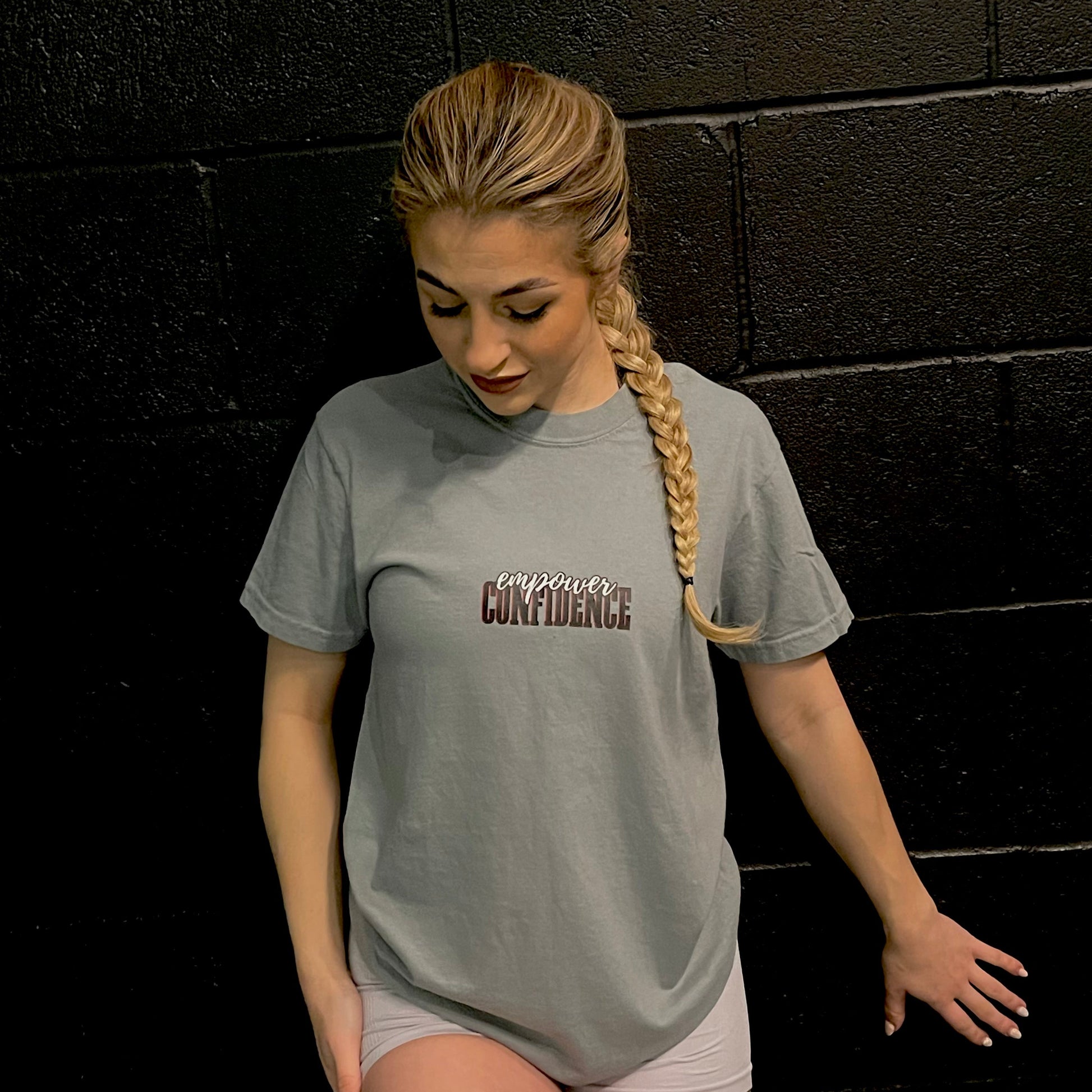 Chase Results clothing brand, based in a gym in Norfolk Norwich, features a confident girl standing in front of a dark backdrop, wearing the Empower Confidence t-shirt. The t-shirt’s bold design highlights her self-assurance, capturing a moment of strength and empowerment as she embraces her reflection with confidence and style.