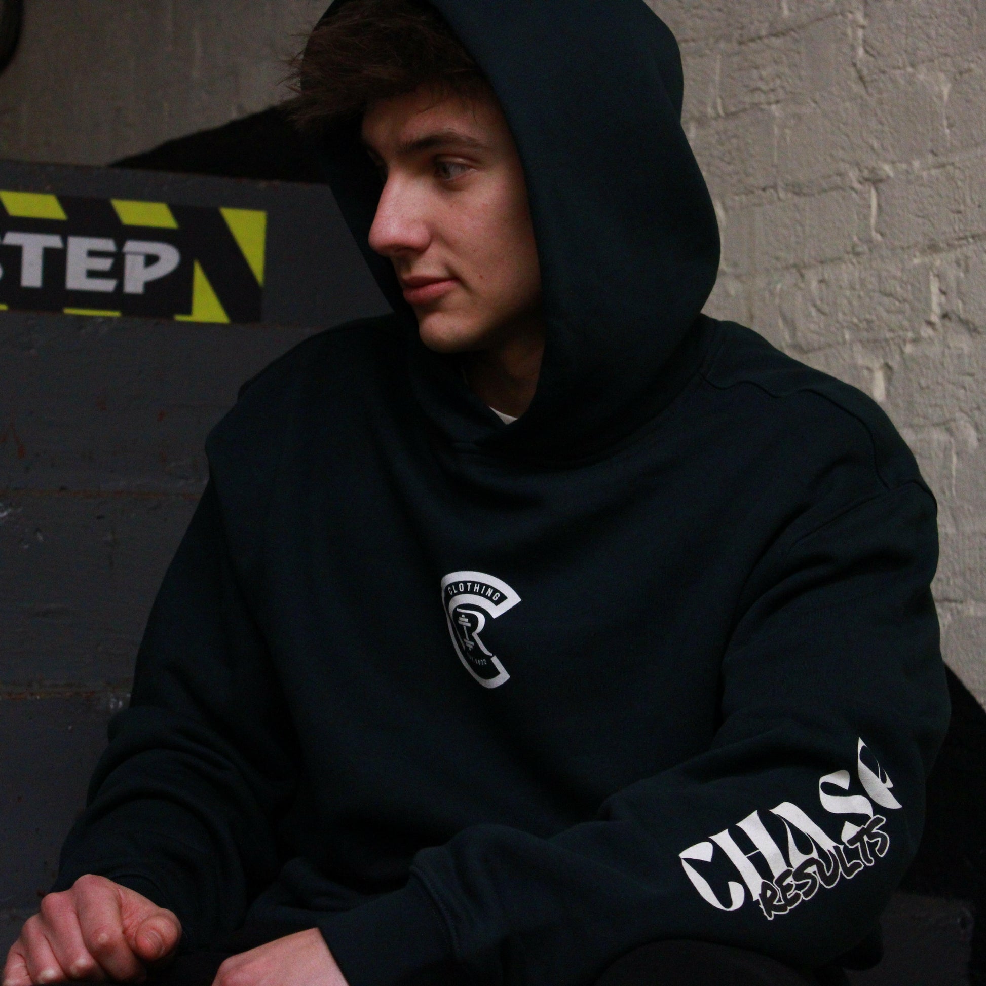 In a dimly lit Norfolk stairwell, a person is seen with their head slightly down and to the side, They wear the Chase Results oversized hoodie by Chase Results Clothing, its bold graphic design enhancing the scenes artistic feel over just any other backdrop. Perfect for those gym lovers, It's Gym wear you can wear anywhere.