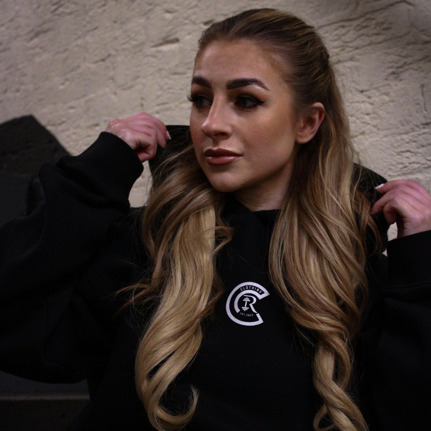 With long, wavy hair, a person pulls up the hood of their limited edition Empower Confidence Black Hoodie by Chase Results Clothing against a textured gray and black wall backdrop, casting soft shadows on their face and exuding empowerment.