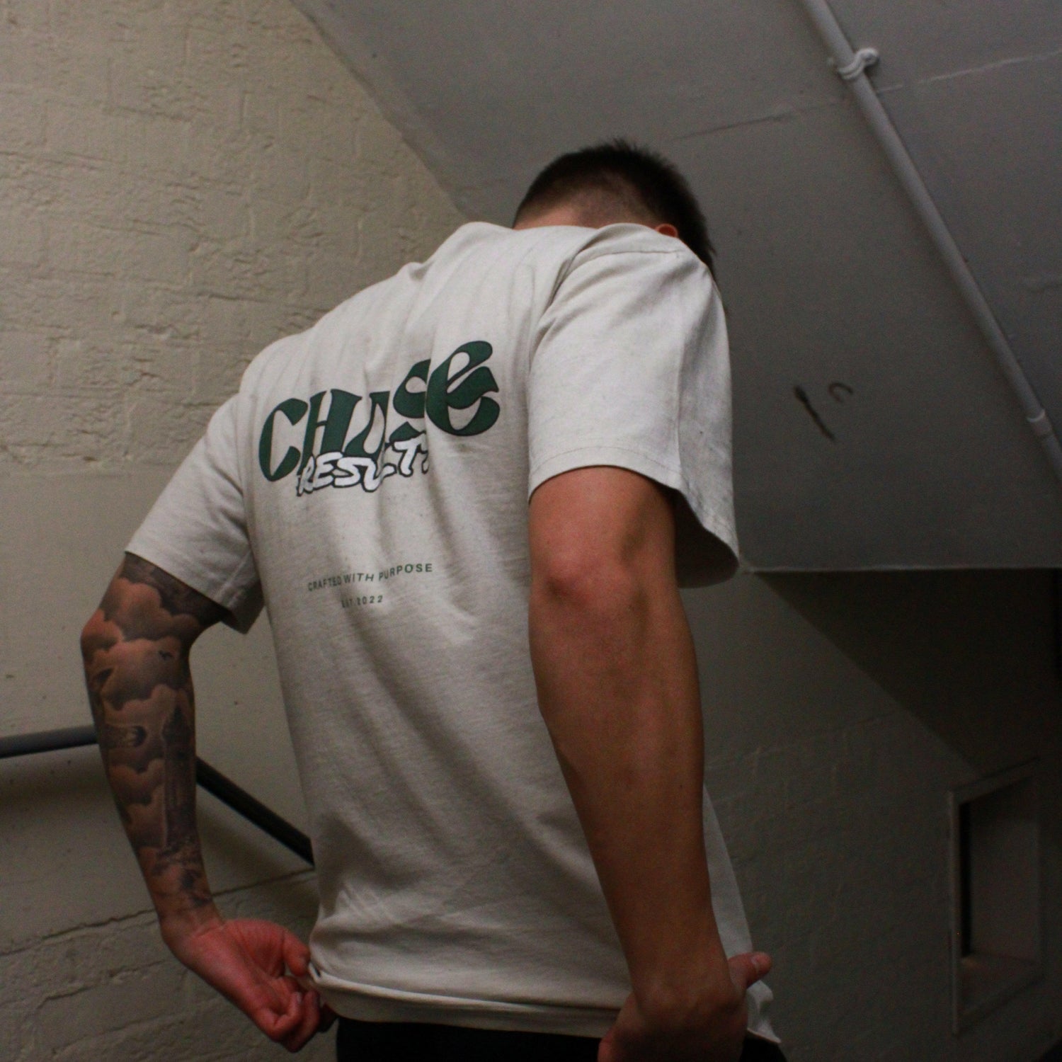 In a dimly lit Norfolk stairwell, a person is seen with their head slightly down and to the side, revealing a tattoo on their left arm. They wear the Chase Results Oversized Pump by Chase Results Clothing, its bold graphic design enhancing the scenes artistic feel over just another gym backdrop.