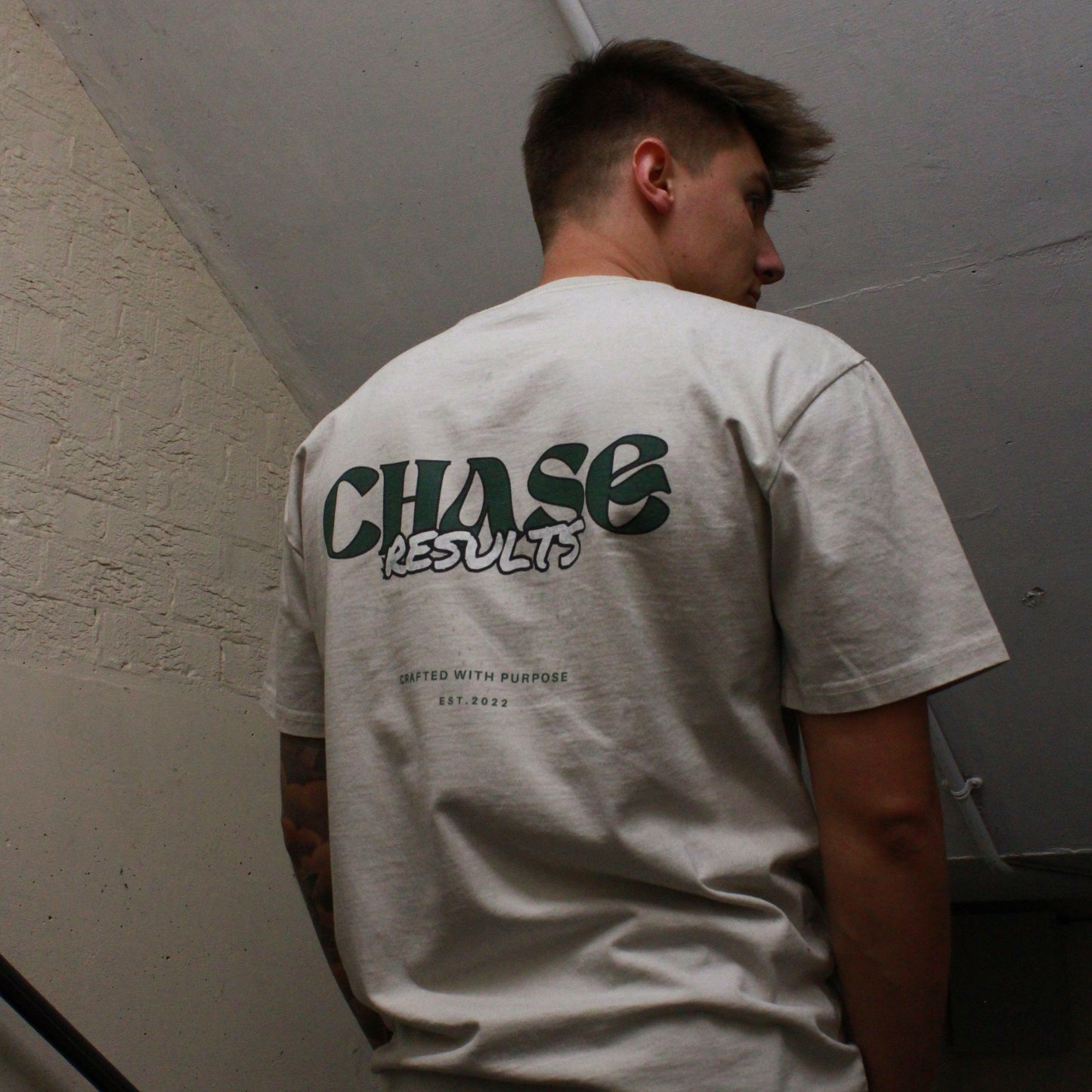 In a dimly lit Norfolk stairwell, a person is seen with their head slightly down and to the side, revealing a tattoo on their left arm. They wear the Chase Results Oversized Pump by Chase Results Clothing, its bold graphic design enhancing the scenes artistic feel over just another gym backdrop.