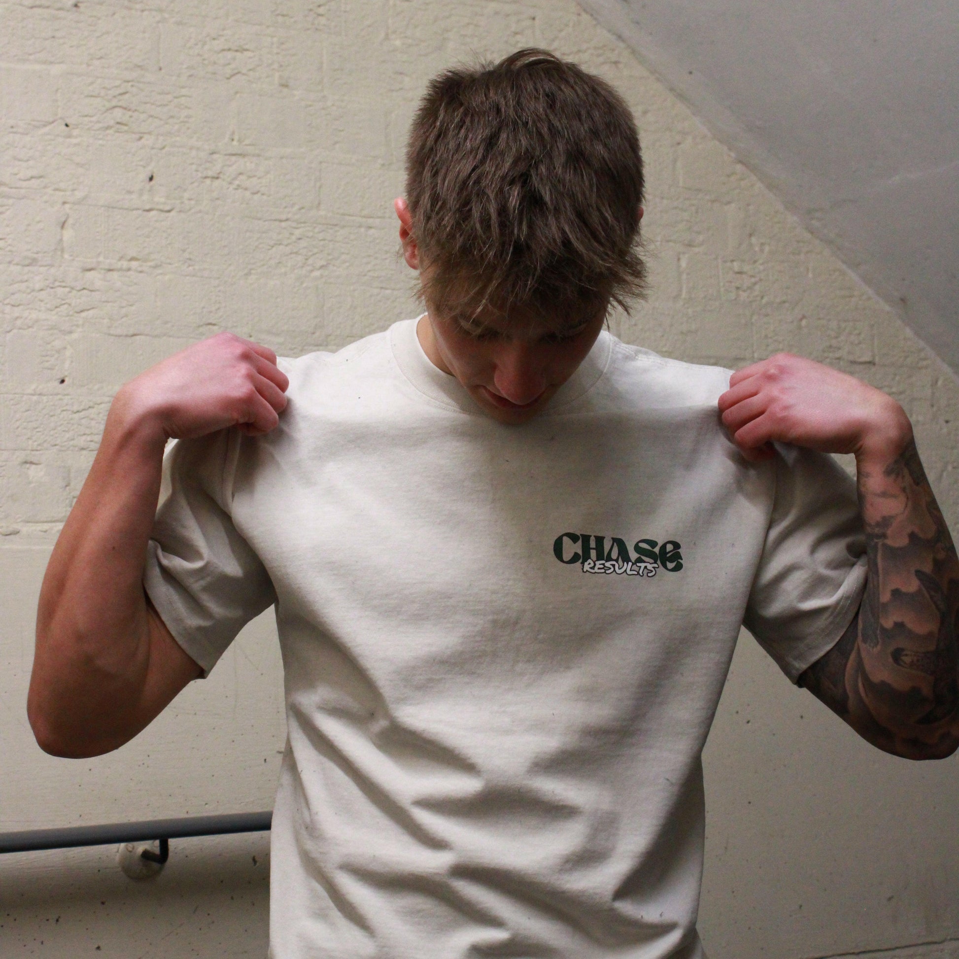 In a dimly lit Norfolk stairwell, a person is seen with their head slightly down and to the side, revealing a tattoo on their left arm. They wear the Chase Results Oversized Pump by Chase Results Clothing, its bold graphic design enhancing the scenes artistic feel over just another gym backdrop.