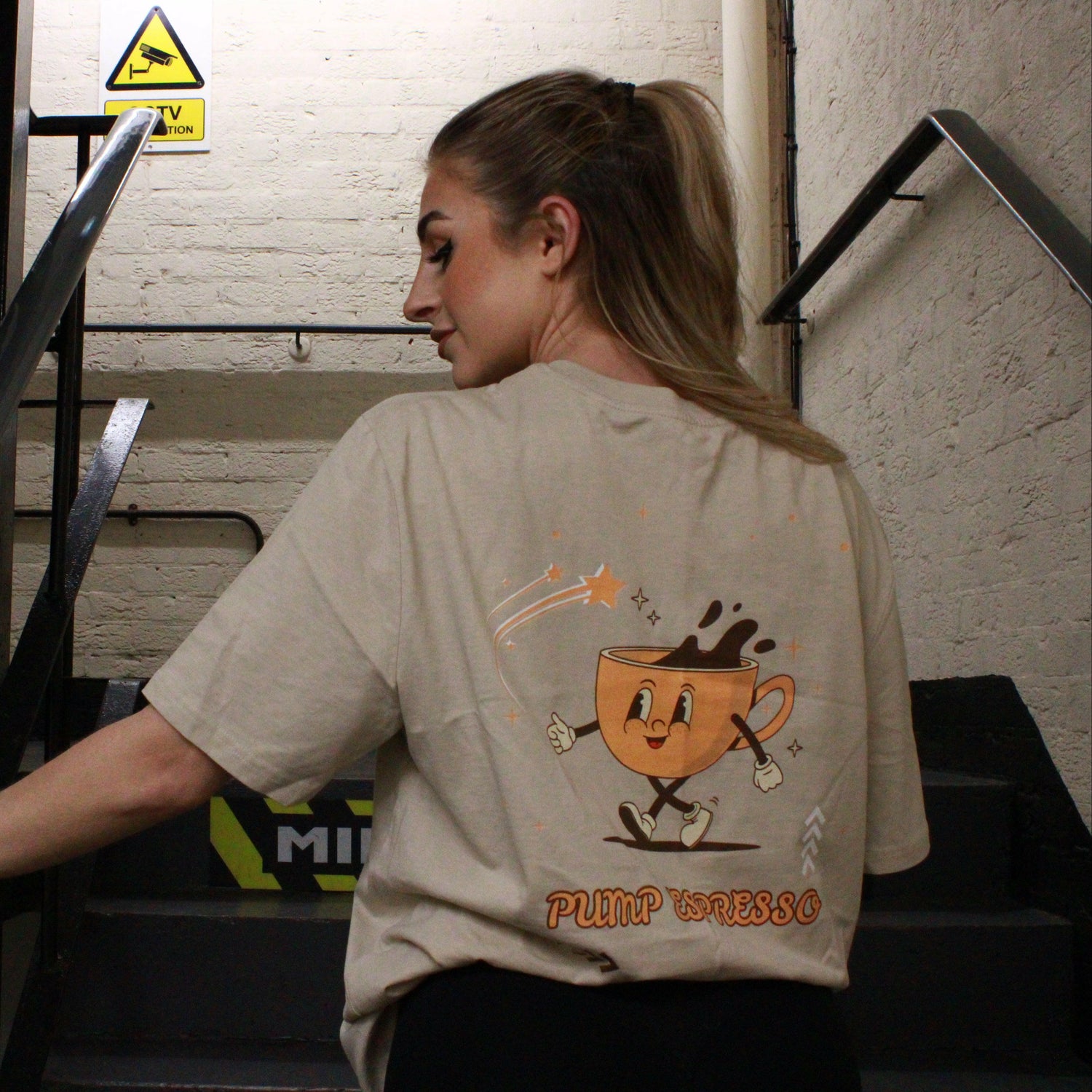 A woman descends the stairs at Great Yarmouth wearing a beige Pump Espresso T-Shirt by Chase Results Clothing, featuring a cartoon coffee cup character. Metal railings frame her path, while a caution sign above ensures safety along her way.