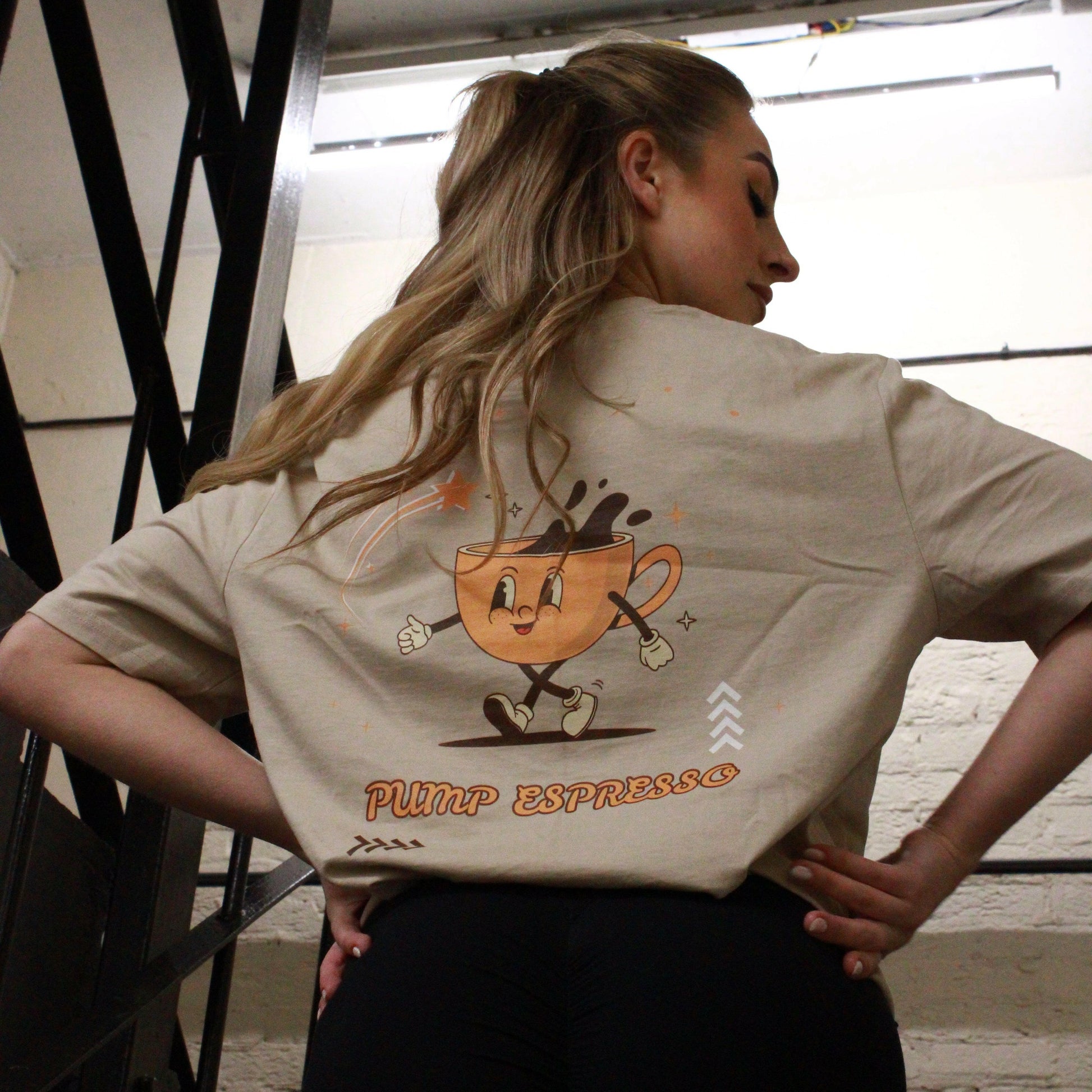 A woman descends the stairs at Great Yarmouth wearing a beige Pump Espresso T-Shirt by Chase Results Clothing, featuring a cartoon coffee cup character. Metal railings frame her path, while a caution sign above ensures safety along her way.