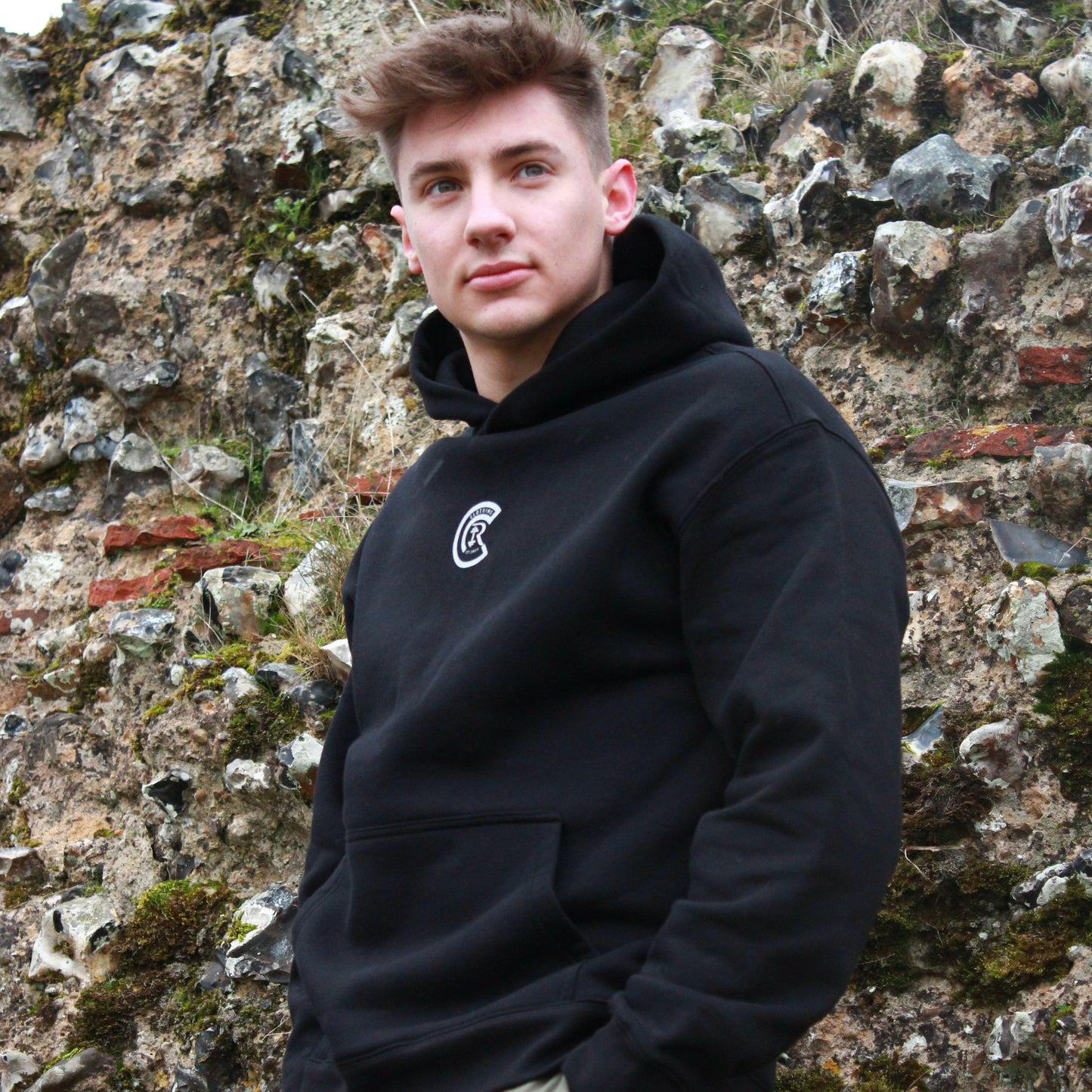 A person with brown hair stands against a mossy stone wall in Norwich, wearing Chase Results Clothing's Basic Black hoodie and Green cargo trousers. Gazing thoughtfully to the side as if contemplating their next move in their journey.