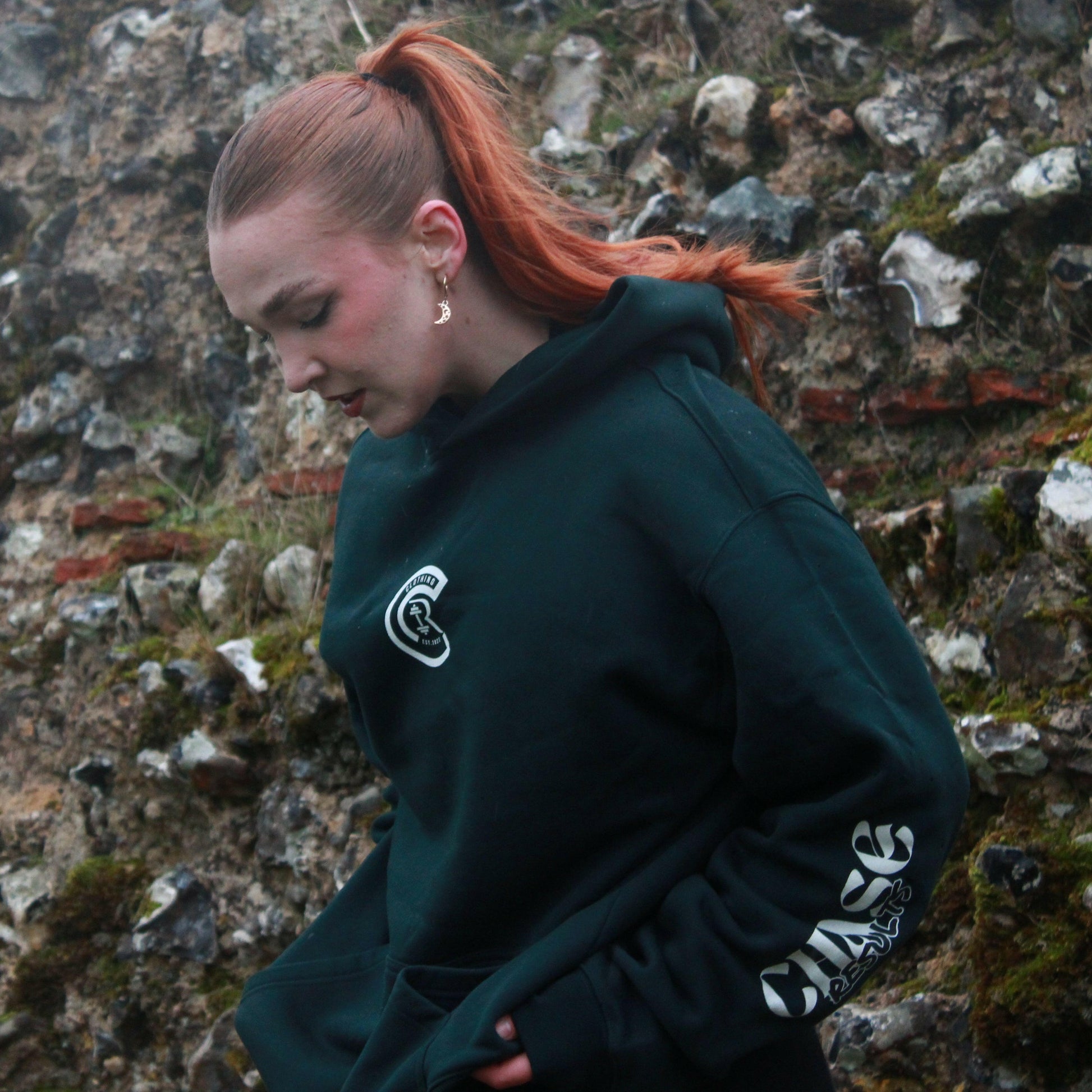 A person in a Chase Results Oversized Hoodie stands against a textured brick wall. The dark green hoodie, ideal for the gym, features CHASE RESULTS boldly on the back with Crafted with Purpose and Est. 2022. Adjust your hood with purpose whether in Norwich or Great Yarmouth.