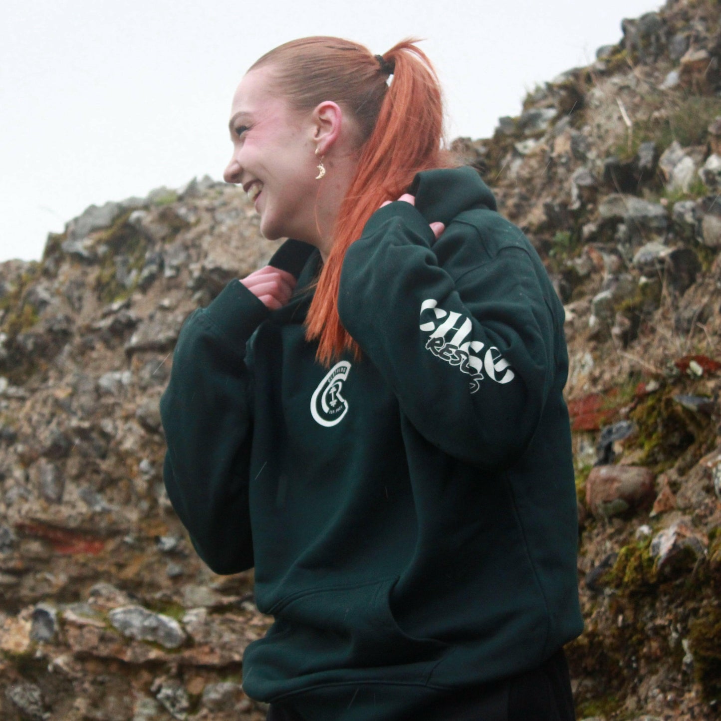 A person in a Chase Results Oversized Hoodie stands against a textured brick wall. The dark green hoodie, ideal for the gym, features CHASE RESULTS boldly on the back with Crafted with Purpose and Est. 2022. Adjust your hood with purpose whether in Norwich or Great Yarmouth.