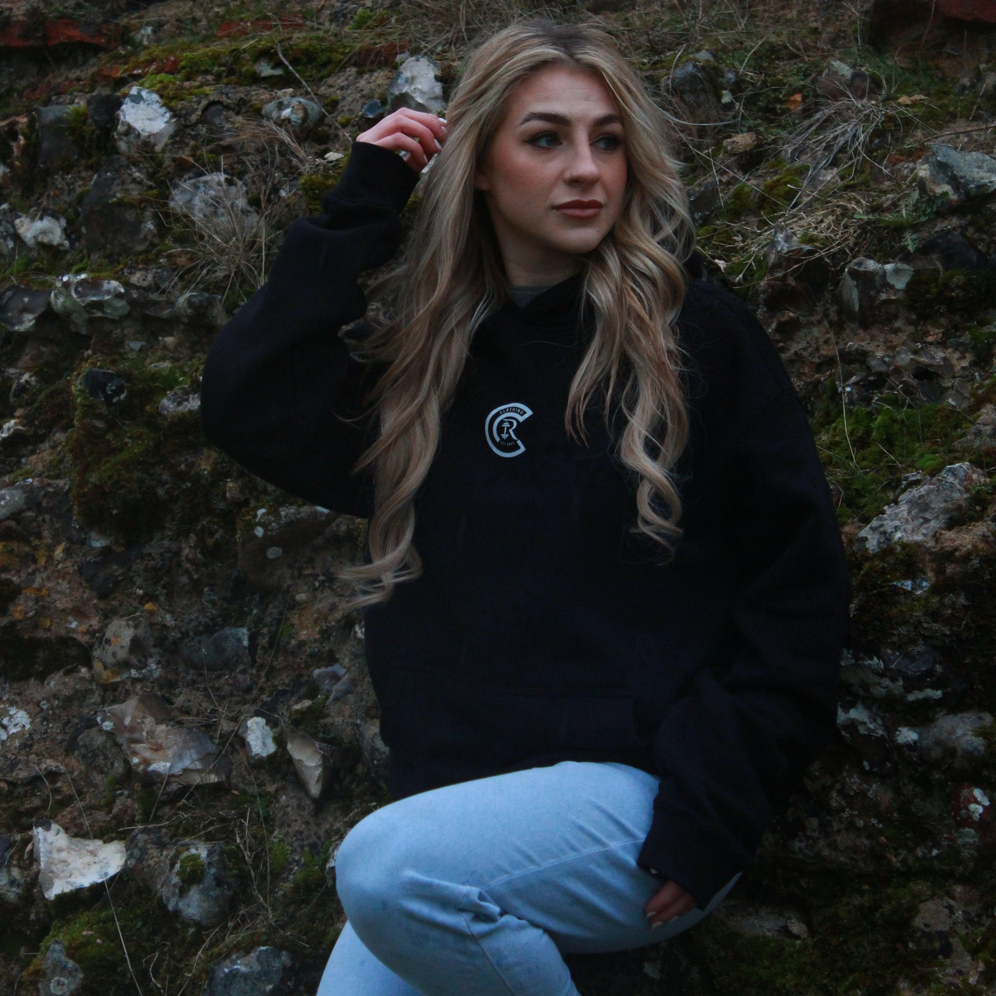 A person with long blonde hair sits against a mossy stone wall in Norwich, wearing Chase Results Clothings Basic Black hoodie and light blue jeans, gazing thoughtfully to the side as if contemplating their next move in their journey.