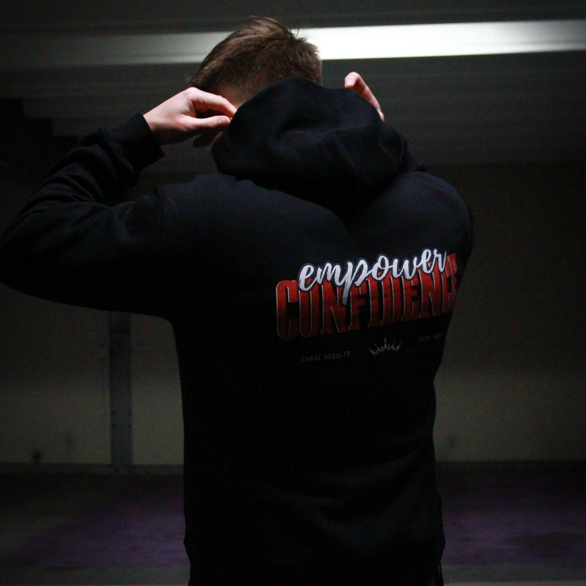 a person pulls up the hood of their limited edition Empower Confidence Black Hoodie by Chase Results Clothing against a textured gray and black wall backdrop, casting soft shadows on their face and exuding empowerment.