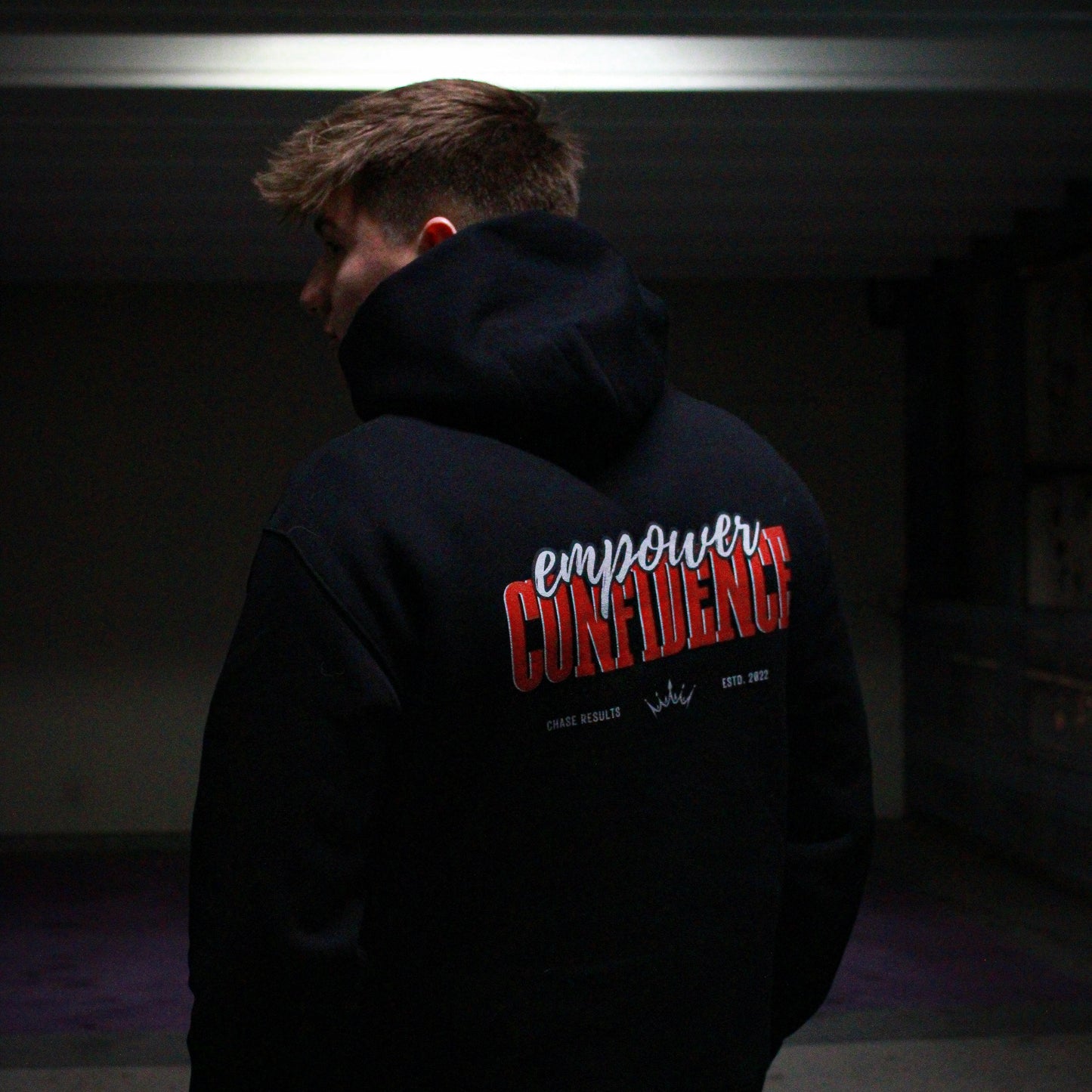 a person hands in their pockets, looking into the distance whilst wearing the limited edition Empower Confidence Black Hoodie by Chase Results Clothing against a textured gray and black wall backdrop, casting soft shadows on their face and exuding empowerment.