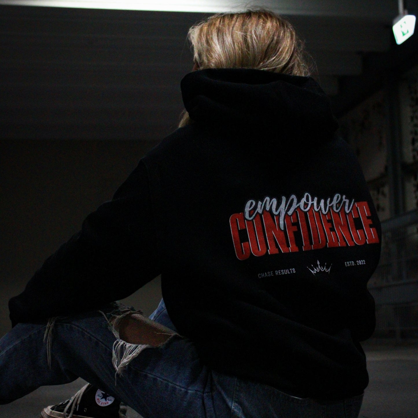 With long, wavy hair, a person pulls up the hood of their limited edition Empower Confidence Black Hoodie by Chase Results Clothing against a textured gray and black wall backdrop, casting soft shadows on their face and exuding empowerment.