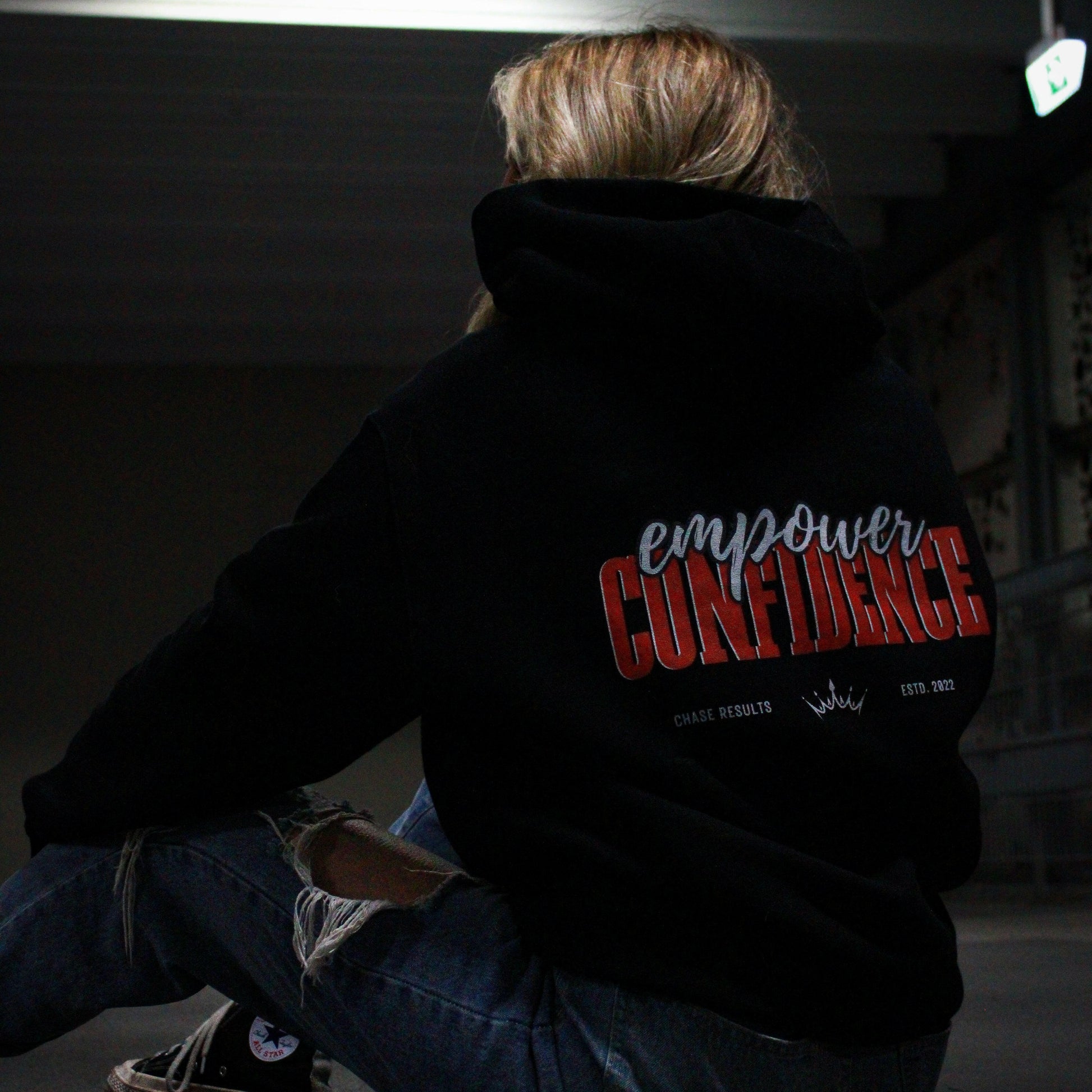 With long, wavy hair, a person pulls up the hood of their limited edition Empower Confidence Black Hoodie by Chase Results Clothing against a textured gray and black wall backdrop, casting soft shadows on their face and exuding empowerment.