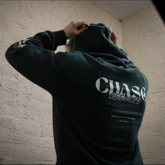 A person in a Chase Results Oversized Hoodie stands against a textured brick wall. The dark green hoodie, ideal for the gym, features CHASE RESULTS boldly on the back with Crafted with Purpose and Est. 2022. Adjust your hood with purpose whether in Norwich or Great Yarmouth.