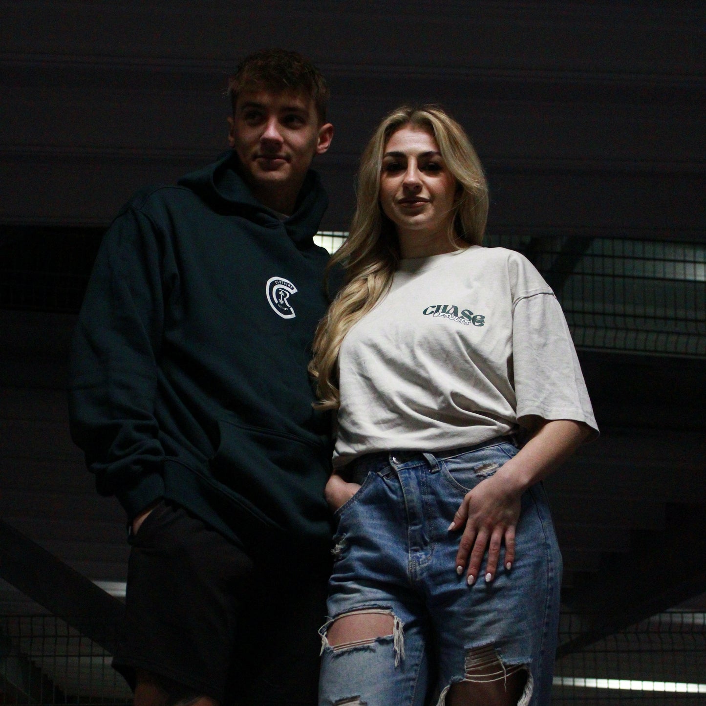 Chase Results clothing brand, based in Norfolk Norwich, features two individuals standing in a dimly lit car park, wearing the brand's stylish apparel. The urban setting highlights the bold and distinctive design of their clothing, perfect for those who want to stand out in any environment. They wear the Chase Results Oversized Pump by Chase Results Clothing, its bold graphic design enhancing the scenes artistic feel over just another gym backdrop.