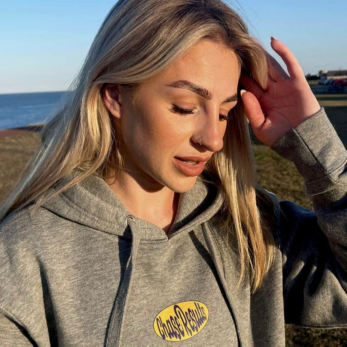 Woman wearing a gray hoodie with a logo, standing outdoors near water.