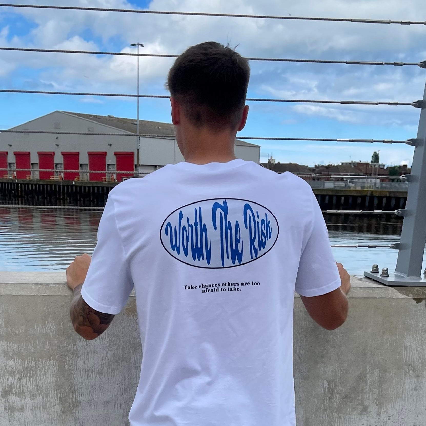 Person wearing a white t-shirt with blue text, standing by a waterfront with a building and sky in the background.