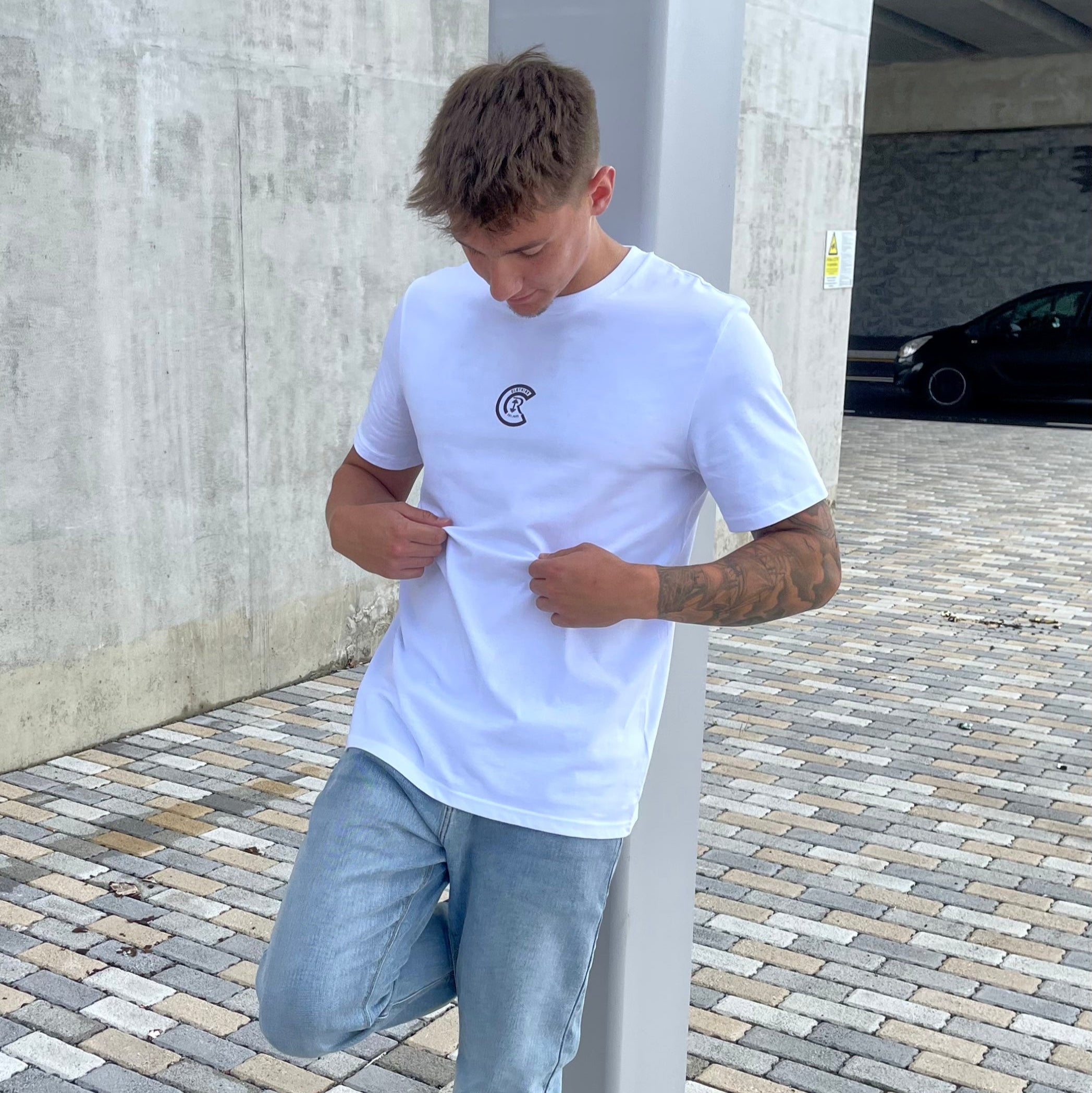 Person wearing a white t-shirt with a logo and light blue jeans standing in an outdoor setting.